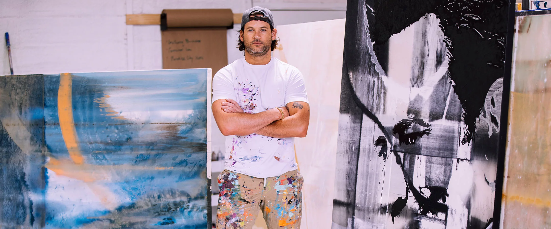Erik Skoldberg standing in his studio in a paint-spattered white shirt, an Urban Collection canvas in blue and gold to his left and a large black-and-white Icon Collection painting to his right