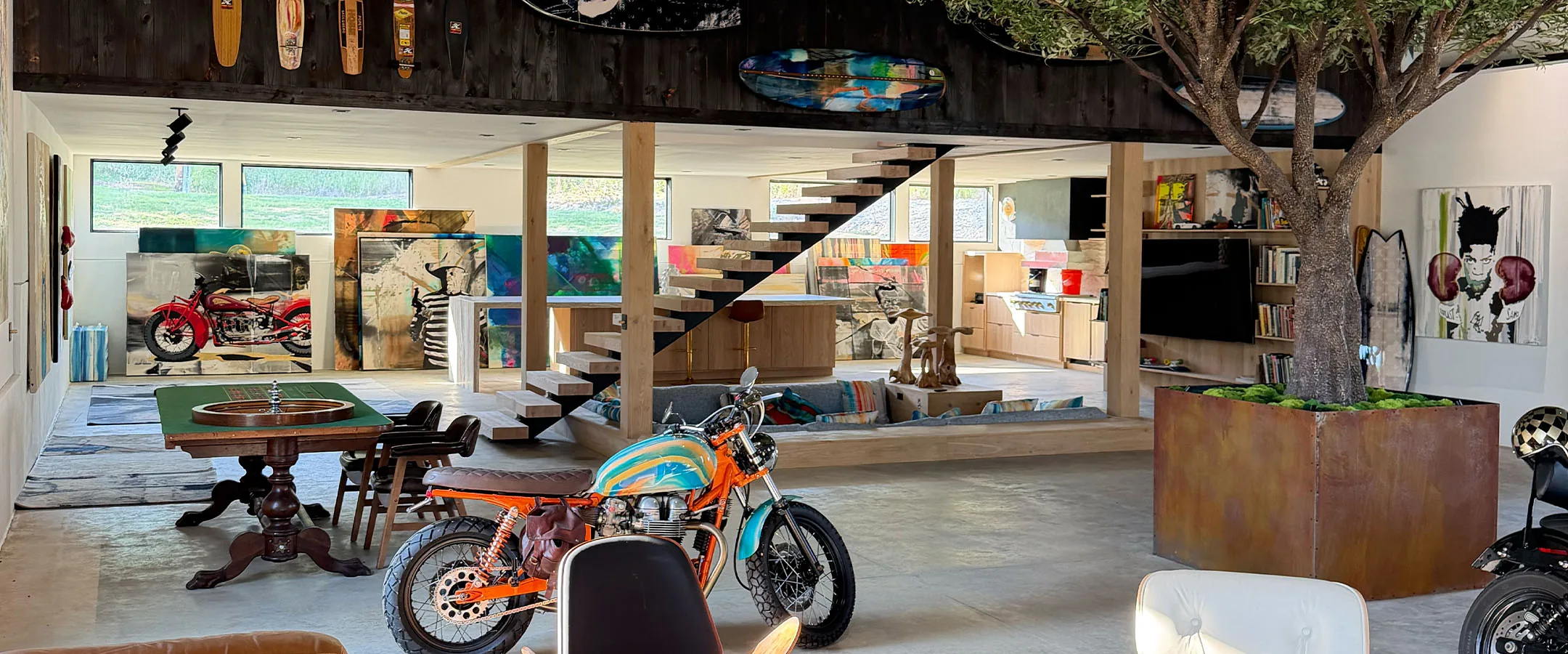 The studio barn in Kingston Springs — painted surfboards and skate decks racked along the dark beam, the floating stair, the olive tree and the motorcycles parked on the floor below