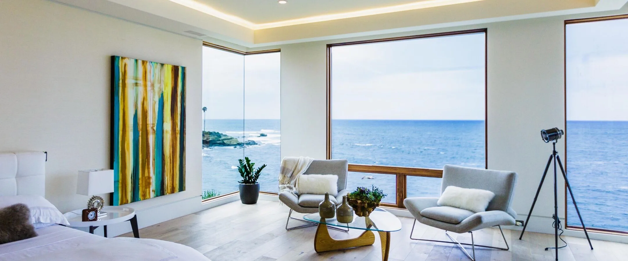 A green, yellow and teal Raw Collection painting by Erik Skoldberg beside the bed in a room with a wall of windows onto the ocean